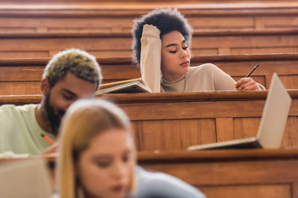 African american student holding pen near laptop and blurred friends in university — Stock Photo