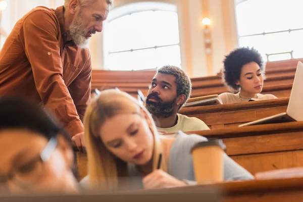 Maturo insegnante in piedi vicino africano americano studente e laptop in auditorium di università — Foto stock