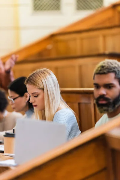 Young student sitting near coffee and interracial friends in university — Stock Photo