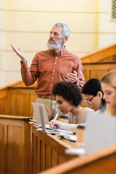 Professeur mature pointant avec la main près des étudiants multiethniques en utilisant des ordinateurs portables à l'université — Photo de stock