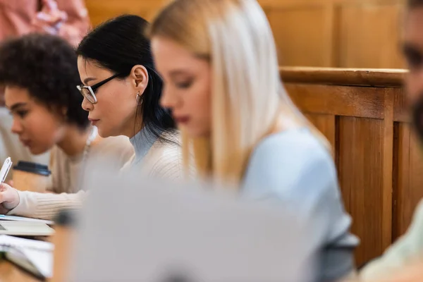 Asian student writing on notebook near blurred multiethnic friends in university auditorium — Stock Photo