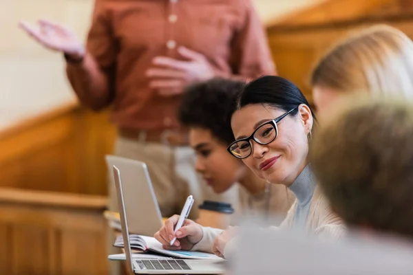 Sourire asiatique étudiant écriture sur notebook près multiethnique amis et ordinateurs portables à l'université — Photo de stock