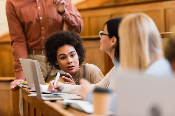 Étudiant afro-américain regardant des amis flous près des ordinateurs portables et professeur à l'université — Photo de stock