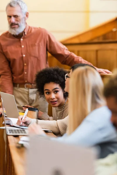 Estudiante afroamericano hablando con amigos cerca de profesor borroso y computadoras portátiles en la universidad - foto de stock