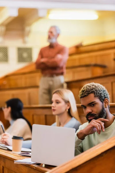 Étudiant afro-américain regardant ordinateur portable près des amis flous et professeur à l'université — Photo de stock