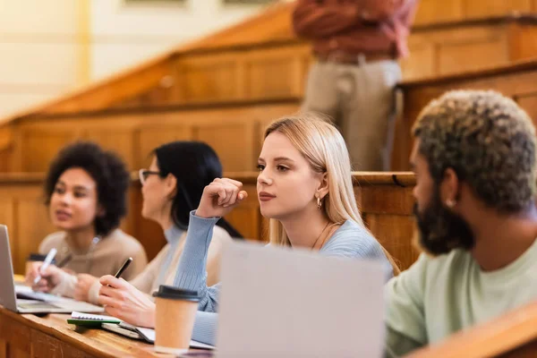 Joven estudiante sentado cerca de amigos multiétnicos y cuadernos en la universidad - foto de stock