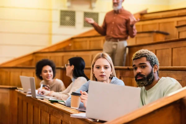 Estudiantes multiétnicos mirando el portátil cerca de amigos borrosos y profesor en la universidad - foto de stock