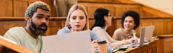 Interracial students using laptop near coffee to go in university, banner — Stock Photo