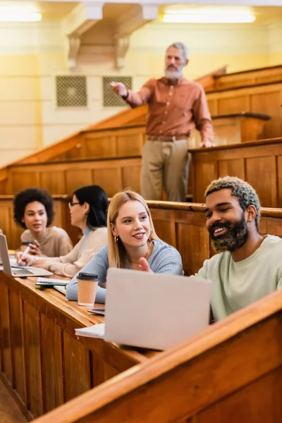 Lächelnde multiethnische Studenten mit Laptop in der Nähe von Kaffee und Freunden an der Universität — Stockfoto