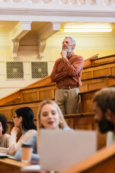 Pensive teacher standing near blurred multiethnic students in university — Stock Photo