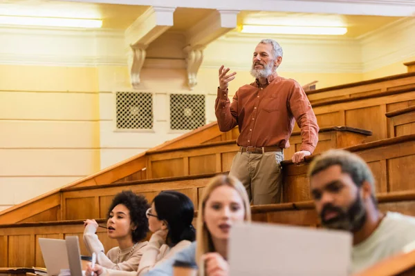 Maduro professor falando perto de alunos inter-raciais turvos com laptops na universidade — Fotografia de Stock