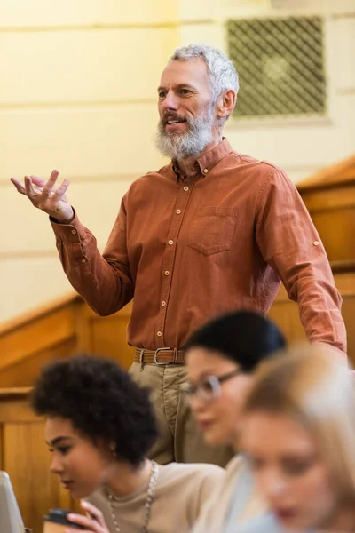 Sonriente profesor hablando cerca borrosa interracial estudiantes en la universidad - foto de stock