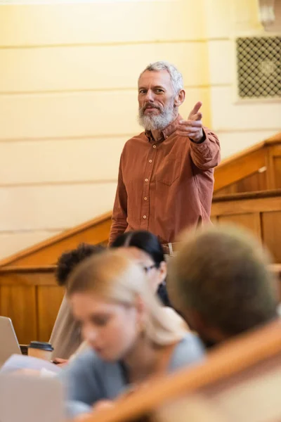 Professor de meia-idade apontando com a mão perto de estudantes multiétnicos e laptops na universidade — Fotografia de Stock