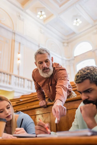 Professeur d'âge moyen avec de la craie parlant à des étudiants multiethniques avec des cahiers à l'université — Photo de stock