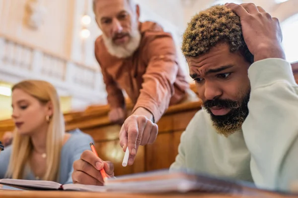 Stressed african american student looking at notebook near blurred teacher and friend in university — Stock Photo