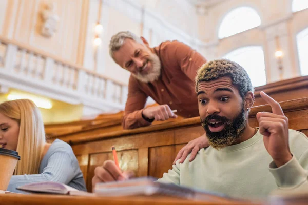 Aufgeregte afrikanisch-amerikanische Studentin schreibt auf Notizbuch neben verschwommenem Lehrer in der Universität — Stockfoto