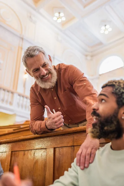 Profesora sonriente con tiza mostrando como gesto cerca de estudiante afroamericano en la universidad - foto de stock