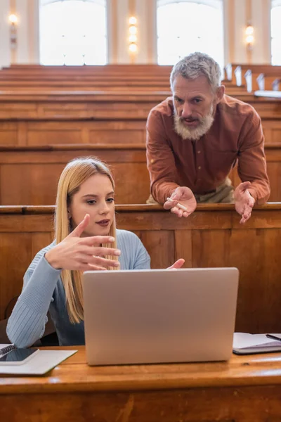 Mature teacher with chalk talking to student near notebooks and devices in university — Stock Photo