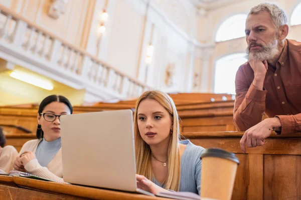 Teacher looking at laptop near interracial students in university — Stock Photo