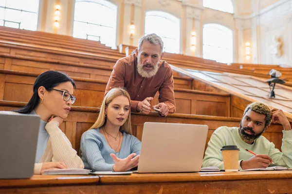 Estudiantes multiculturales mirando el portátil cerca del profesor con tiza en la universidad - foto de stock