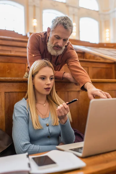 Joven estudiante apuntando a la computadora portátil borrosa cerca del profesor en la universidad - foto de stock