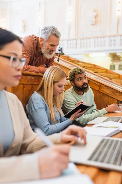 Professora sorrindo olhando para estudantes inter-raciais com cadernos e laptops na universidade — Fotografia de Stock