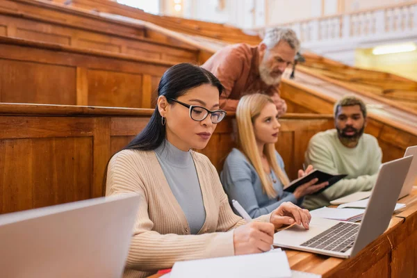 Reife asiatische Studenten schreiben auf Notebook in der Nähe von Laptop, verschwommen Freunde und Lehrer in der Universität — Stockfoto