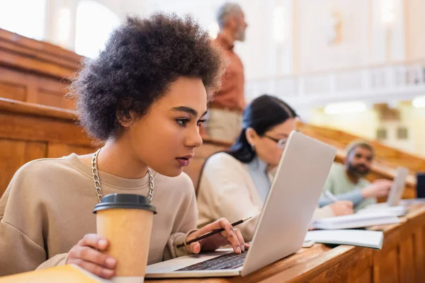 Young african american student using laptop and holding coffee to go near friends in university — Stock Photo