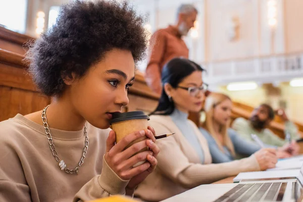 African american student holding coffee to go near laptop in university — Stock Photo