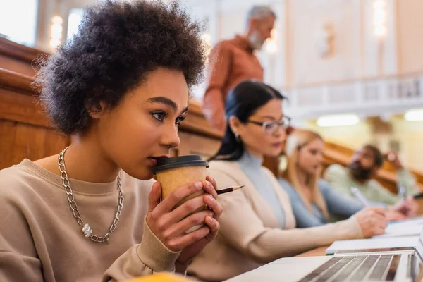 Estudiante afroamericano sosteniendo café para ir cerca de portátil en la universidad - foto de stock