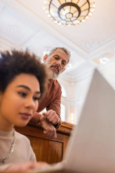 Mature teacher looking at blurred african american student with laptop in university — Stock Photo