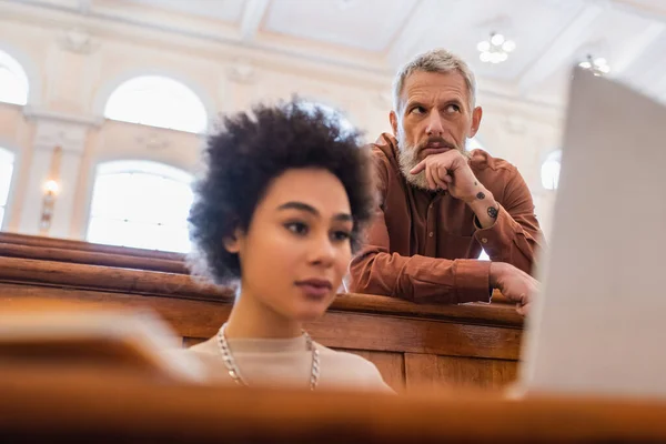 Pessimistischer Lehrer in der Nähe verschwommener afroamerikanischer Student mit Laptop in Universität — Stockfoto