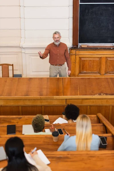 Professeur mature pointant avec la main près des étudiants interraciaux écrivant sur des cahiers à l'université — Photo de stock