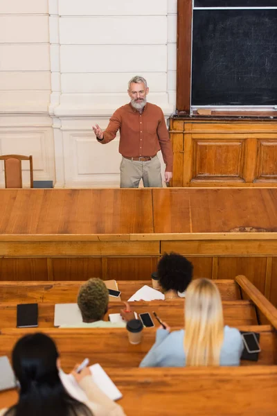 Maturo insegnante che indica con mano vicino a studenti interrazziale scrivere su taccuini in università — Foto stock