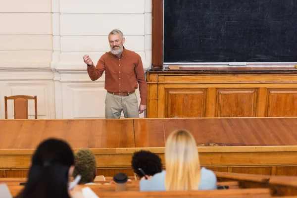Sonriente profesor hablando durante conferencia cerca borrosa interracial estudiantes en la universidad - foto de stock