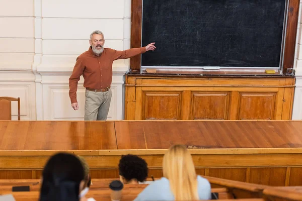 Insegnante indicando lavagna vicino sfocato interrazziale studenti in università — Foto stock