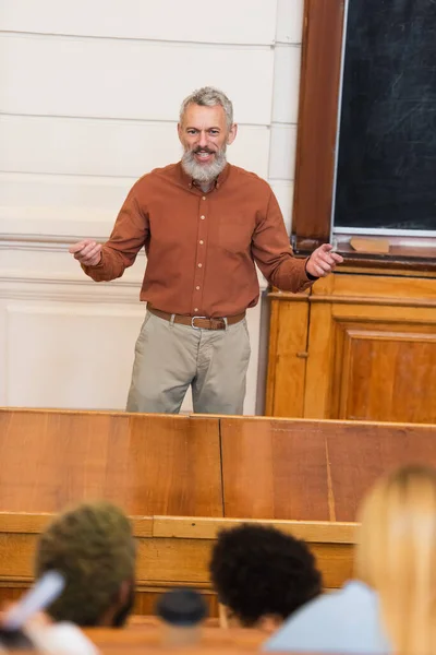 Insegnante sorridente che parla vicino alla lavagna e studenti multietnici offuscati all'università — Foto stock