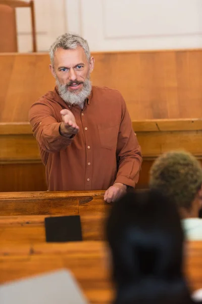 Profesor de mediana edad señalando con la mano al estudiante borroso en la universidad - foto de stock