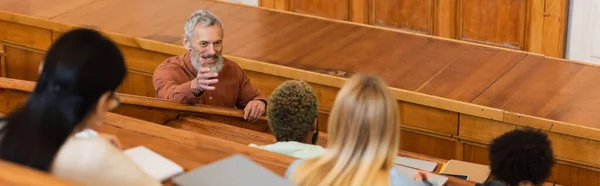 Professeure souriante parlant à des étudiants interraciaux avec des cahiers à l'université, bannière — Photo de stock