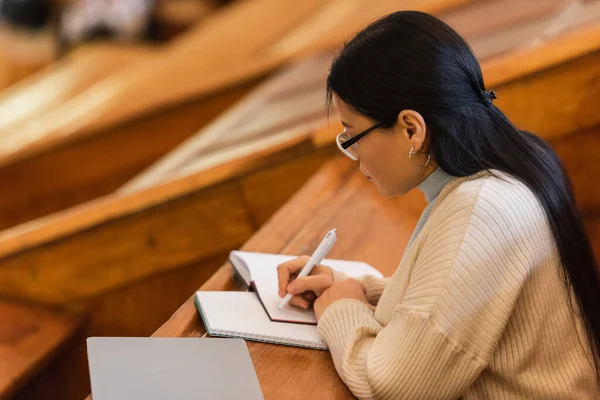 Side view of asian student writing on notebook near laptop in university — Stock Photo