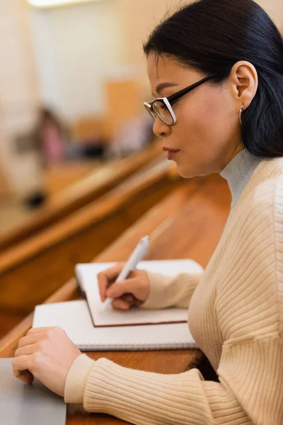 Seitenansicht einer asiatischen Studentin, die in der Nähe ihres Laptops auf einem verschwommenen Notizbuch schreibt — Stockfoto