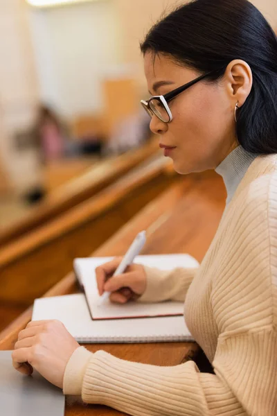 Side view of asian student writing on blurred notebook near laptop in university — Stock Photo