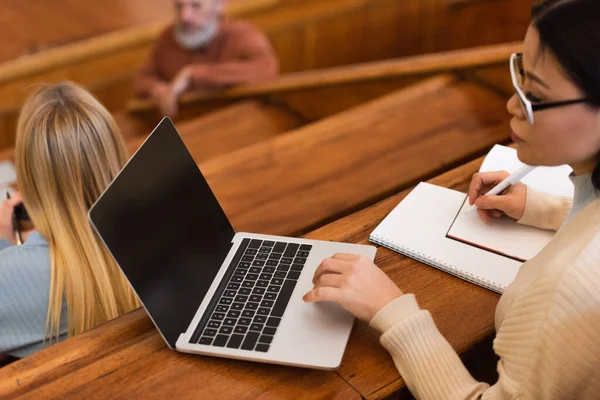 Blurred asian student using laptop and writing on notebook in university — Stock Photo