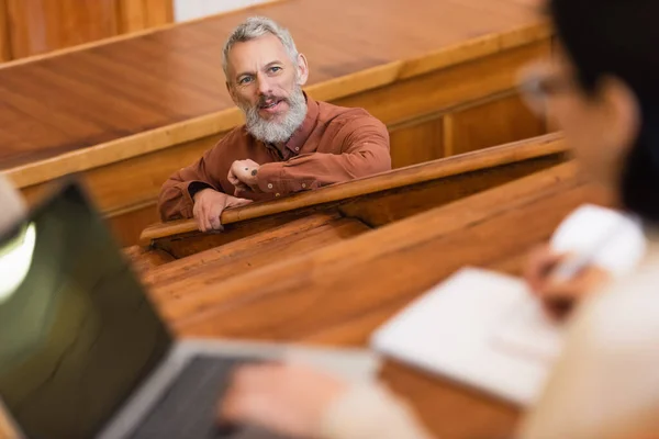 Enseignant d'âge moyen regardant flou étudiant avec ordinateur portable pendant la conférence à l'université — Photo de stock