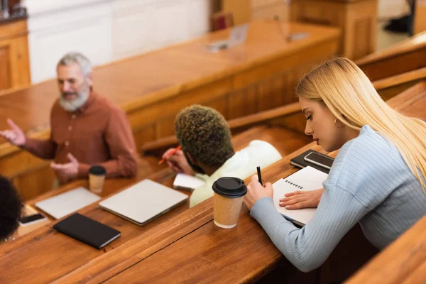 Student writing on notebook near coffee to go and blurred interracial friends in university — Stock Photo