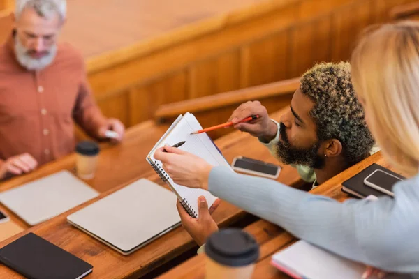 Estudantes inter-raciais segurando notebook perto de dispositivos e professor turvo na universidade — Fotografia de Stock