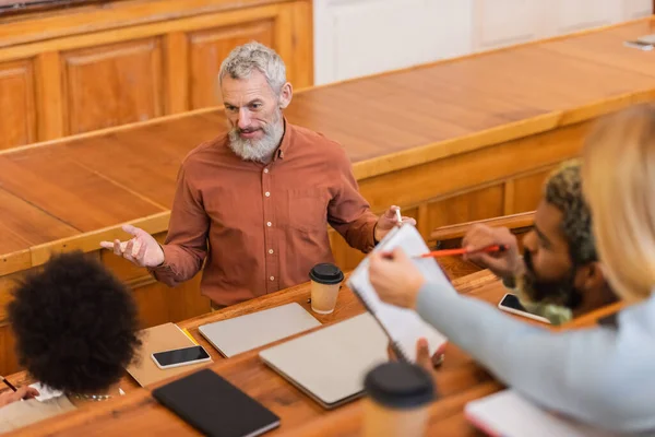 Teacher with chalk talking near multiethnic students and devices in university — Stock Photo
