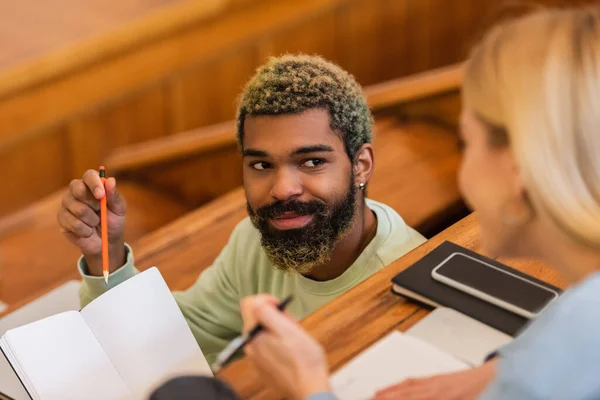Estudante afro-americano sorrindo apontando para notebook perto de amigo borrado na universidade — Fotografia de Stock