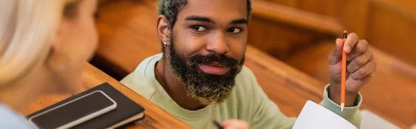 Studente afroamericano sorridente che indica il taccuino vicino ad un amico offuscato e al cellulare all'università, striscione — Foto stock