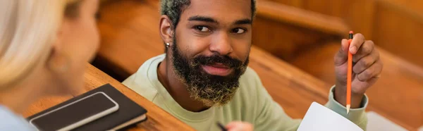 Smiling african american student pointing at notebook near blurred friend and cellphone in university, banner — Stock Photo